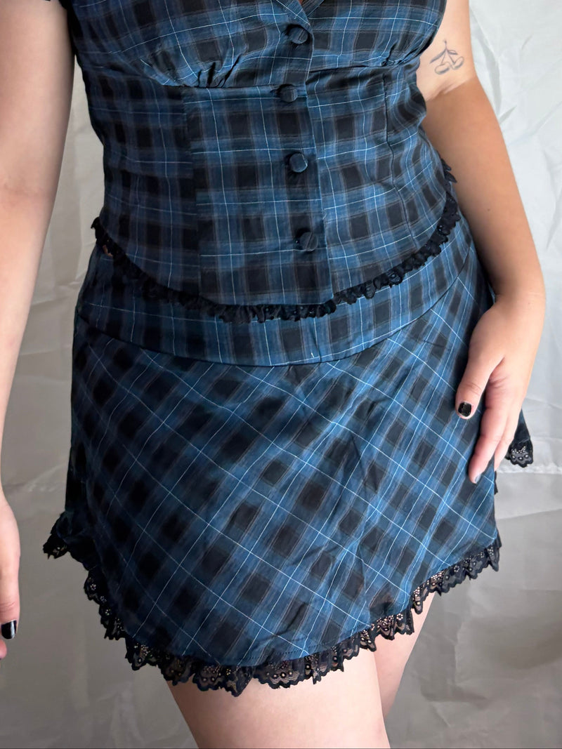 A woman modeling a short-sleeve plaid mini dress with a button-front bodice, ruffled trim, and lace hem against a wrinkled white fabric backdrop.